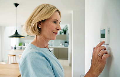 A mature, smiling woman adjusts a thermostat