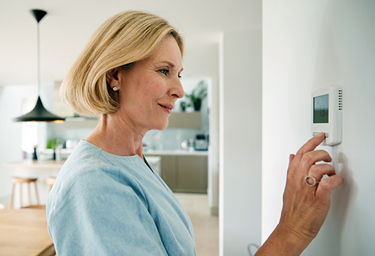 A mature, smiling woman adjusts a thermostat