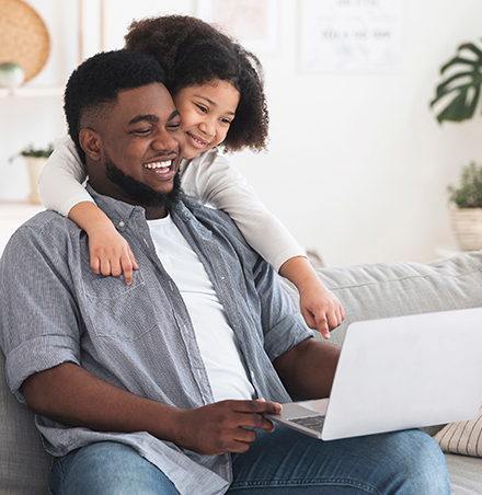 A father smiling and holding a laptop while his daughter is smiling and pointing to the screen